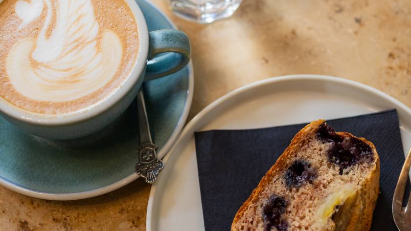 Ein Stück hausgemachtes Banana Bread mit Cappuccino und Wasserglas auf einem Tisch im MANANA cafẽ in Wien Währing.