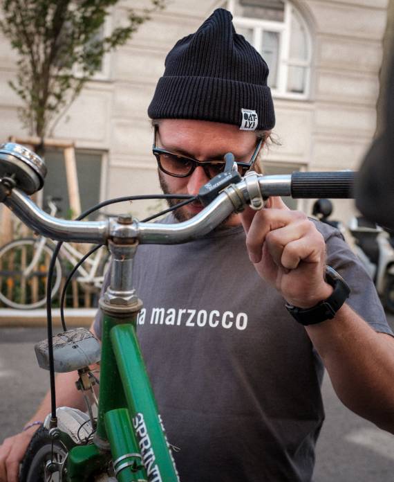 Dirk steht vor dem MANANA café, schraubt an einem Puch-Fahrrad, trägt eine Oatly-Haube und ein La Marzocco Shirt